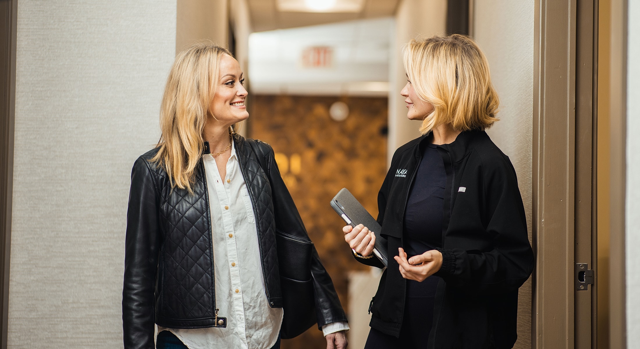Two women talking in a hallway setting.